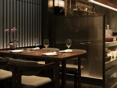 A dimly lit restaurant dining area with a table, chairs, and counter, featuring a combination of wood and metal elements, wine glasses, and decorative items