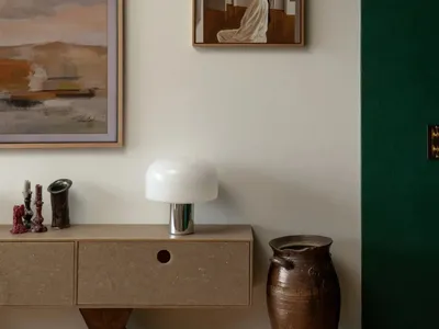 A well-decorated living room with a white wall featuring a variety of artwork, a vase on a table, and a lamp, creating an inviting atmosphere