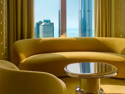 A well-lit living room with a couch and dining table, featuring a large window with a city view, wood furniture, and glass accents