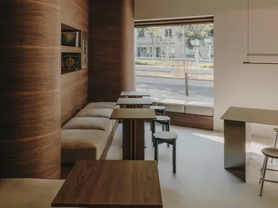 A modern restaurant interior with a large wooden dining area, featuring multiple chairs, benches, and a wooden table, set against a well-lit background with a bar in the distance