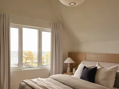 A calm bedroom with a white bed, a window, and a table lamp, surrounded by a white color scheme and natural light