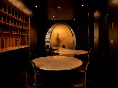 A cozy restaurant dining area with a large round table, wine glasses, and a server, surrounded by wood and glass elements, with soft and warm lighting