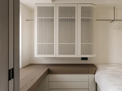 A minimalist bedroom with a wooden desk, white cabinet, and a bed with a white comforter and pillows, lit by natural light
