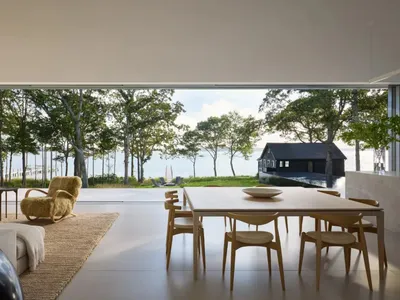 A well-lit living room with a dining table, couch, and chair, surrounded by a large window showcasing a beautiful water view