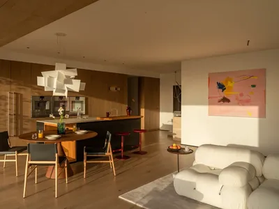 Spacious living room with a wooden floor, a dining table, kitchen area, and natural light from windows, decorated with a vase, potted plant, and bowl