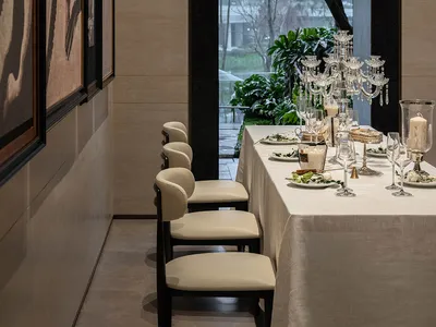Dining room with a large table set for a meal, surrounded by potted plants and a large window allowing natural light