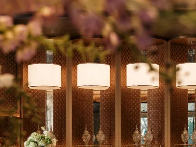 A modern bar with a marble counter, stools, and a vase of flowers, decorated with bottles and cups, and two potted plants, under soft and warm lighting