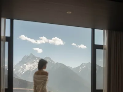 A woman stands in a living area with a couch and dining table, looking out at the mountains through a large window, surrounded by a cozy and inviting atmosphere with natural light and a vase with flowers on the table