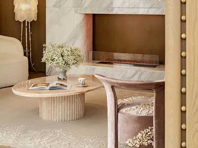 Cozy living room with a pink and white color scheme, featuring a marble fireplace, a couch, and a chair, with a dining table and vase adding to the aesthetic