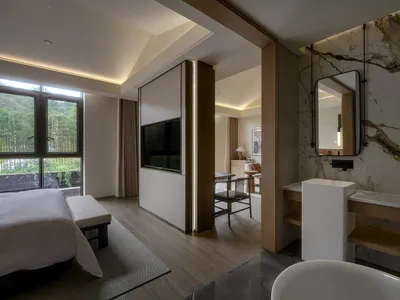 A modern hotel room with a bed, TV, chair, and desk, featuring wooden floors and a marble countertop, in a well-lit and inviting space