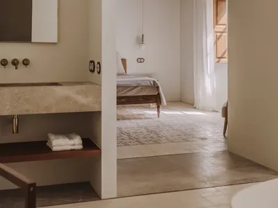 A minimalist bedroom with a bed, a wooden chair, and a white rug, well-lit by natural light from the window