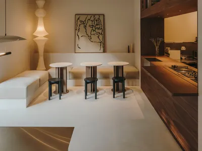 A well-lit living room with a dining area, featuring a dining table, chairs, a couch, bookshelf, vase, and potted plant, in a predominantly white space with a modern appearance