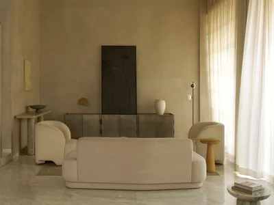 A cozy living room with a beige color scheme, featuring a large couch, a chair, and various decorative pieces, with natural light streaming in through the windows