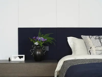 A well-lit bedroom with a bed, nightstand, and vase with flowers, featuring predominantly white materials and a clean appearance