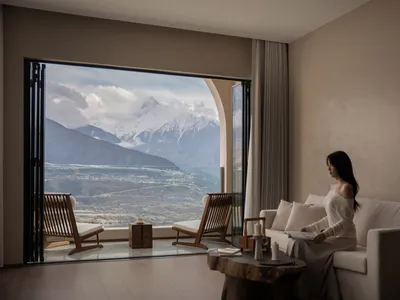 Serene living room with a woman enjoying a mountain view from a large window, surrounded by a couch, chair, dining table, and potted plant
