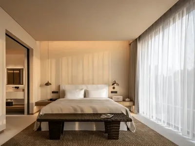 A well-lit hotel bedroom with a bed, bench, and lamp, featuring a white color scheme and natural light from the window