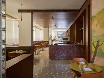 A modern restaurant interior with a dining area featuring a long wooden table and chairs, and a kitchen area with a counter and appliances, lit by soft warm lighting