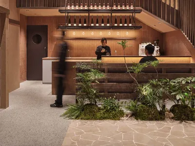 Warm and inviting restaurant interior with a bar, staircase, and potted plants, featuring wood accents and a large fan