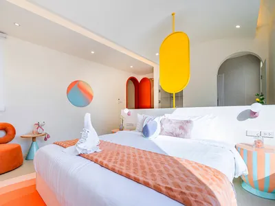 Cozy bedroom with a bed, chair, and yellow and orange color scheme, featuring a white sheet and red and orange checkered blanket, with natural light streaming in through a window