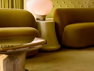 A cozy living room with a green couch and chair, a pink lamp, and a well-lit atmosphere