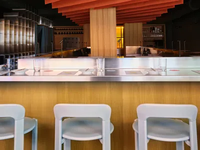 Cozy restaurant interior with a dimly lit bar area featuring a wooden counter and white chairs, surrounded by bottles, cups, and bowls