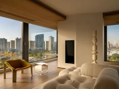 A well-lit living room with a wooden floor, white furniture, and a large window offering a city view, featuring a couch, chair, and TV