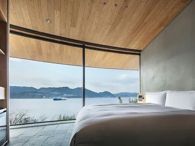A serene bedroom with a large bed, white bedding, and a view of the ocean through a window, bathed in natural light