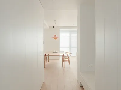 A bright and welcoming room with a wooden floor, a bed on the right, a dining table and chair in the center, and white walls