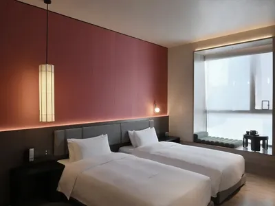 Cozy modern hotel room with two beds, a pink accent wall, and a red wall, featuring white bedding, two lamps, and a comfortable chair near a window with natural light