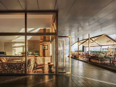 A modern restaurant interior with a wooden floor, dining tables, and chairs, featuring a large window and soft warm lighting