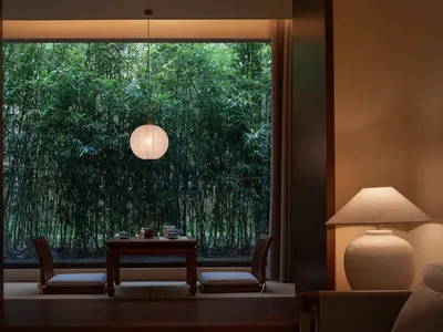 A dining room with a wooden table and chairs, a lamp, and a window with a forest view, creating a warm and inviting atmosphere