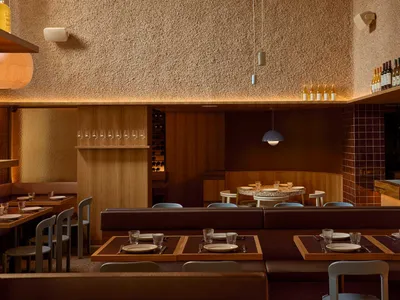 A well-lit restaurant dining area with a long wooden table and comfortable chairs, surrounded by wine glasses, cups, and bowls