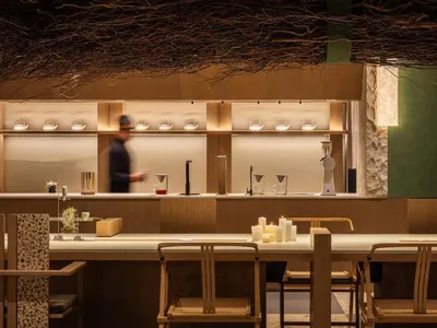 A modern restaurant dining area with a long wooden table, chairs, and vases, set with wine glasses, cups, and bowls, under warm natural light