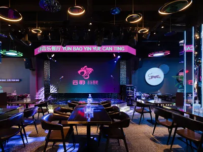 A well-lit restaurant interior with dining tables, chairs, and a large screen, surrounded by bottles and creating a comfortable atmosphere