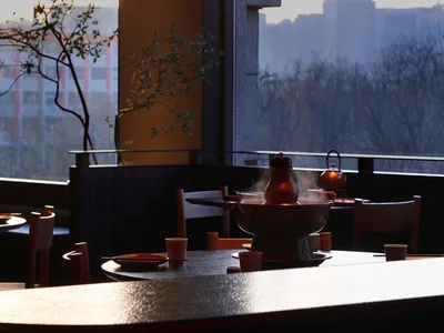 Dining area with a table, chairs, and red accents, set with a bowl and a vase, in a modern restaurant with natural lighting and a warm color palette