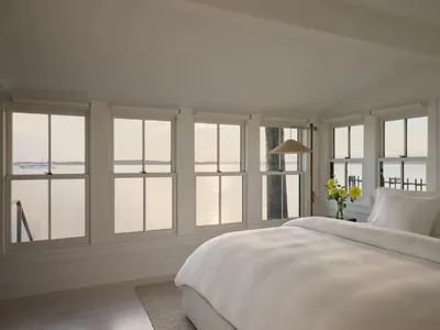 A serene bedroom with a white comforter, yellow flowers, and a wooden floor, lit by natural sunlight through the window