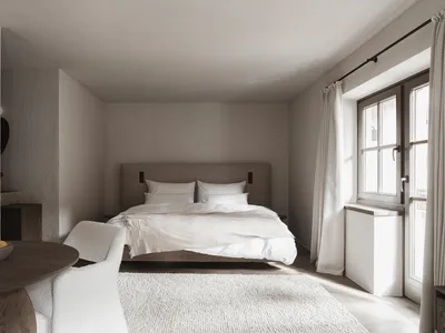 A dimly lit bedroom with a bed and chair, both white, near a window allowing natural light
