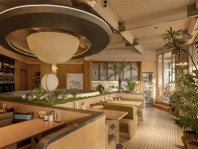 Cozy restaurant interior with a mix of seating options, potted plants, and a circular light fixture, featuring wood and metal elements and soft warm lighting