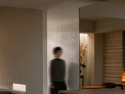 A woman walks through a modern kitchen with a wooden floor and a white wall, featuring a dining table and chair
