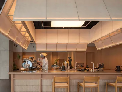 Dimly lit kitchen with a wooden counter, chairs, and dining table, featuring a coffee maker and a cup on the counter
