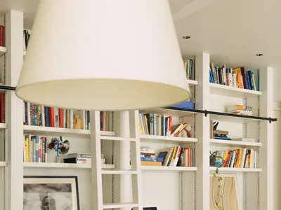 Cozy living room with a white bookshelf, couch, and dining table near a window, featuring a lamp and a comfortable arrangement of books and furniture