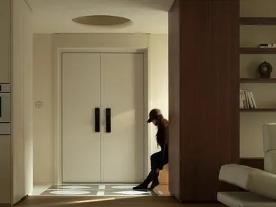 A woman stands in a calm and inviting living space with a wooden door and a white wall, looking through a doorway with a couch visible in the background