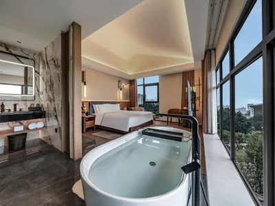 A modern hotel room with a large bathtub, bed, chair, and window, decorated with marble elements and filled with natural light