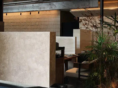 A modern restaurant space with a wooden floor, stone wall, and comfortable seating areas with dining tables and chairs, well-lit with natural light and a potted plant