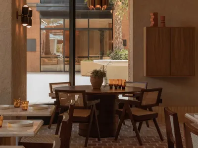 A modern restaurant interior with a dining table, chairs, potted plants, a kitchen with a refrigerator, oven, and sink, and a warm color palette