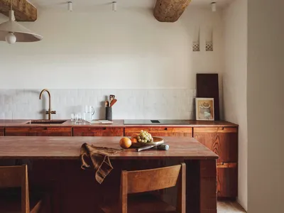 Modern kitchen with a wooden countertop, dining table, and chairs, featuring a sink, refrigerator, and bowl, with a wooden floor and tiled wall, lit by natural sunlight