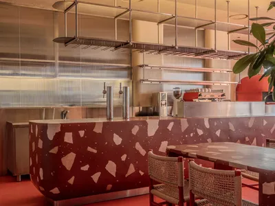 A modern restaurant dining area with a red table and chairs, surrounded by potted plants, warm lighting, and natural light from windows