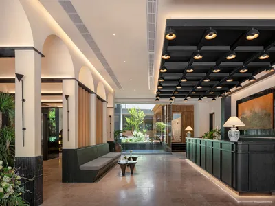 A modern hotel lobby with a large couch, bench, and dining table, surrounded by potted plants and illuminated by natural light