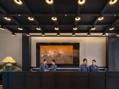 A bright and welcoming hotel lobby with a modern style, featuring a large painting, potted plants, a couch, and a chair, lit by bright lighting
