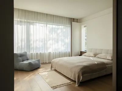 A modern bedroom with a bed, chair, and window, featuring a predominantly white color scheme and natural light from the window